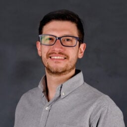 a man with short dark hair, glasses, and a beard is smiling at the camera. he is wearing a light gray button up shirt and is posed against a plain dark gray background.